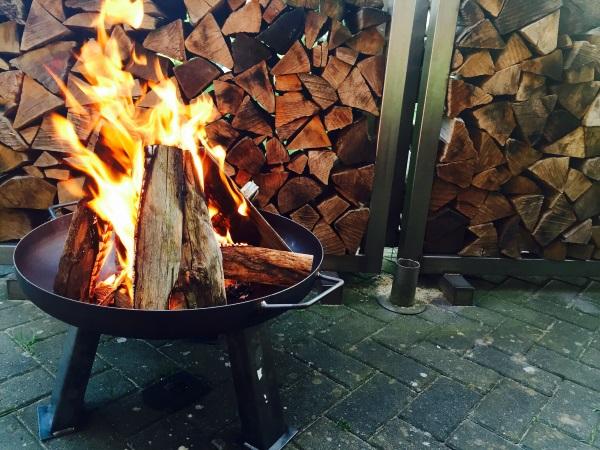 In einer feuerschale brennt ein Feuer. Im Hintergrund ist geschichtetes Holz zu sehen. Feuerschale zum Kochen und Grillen