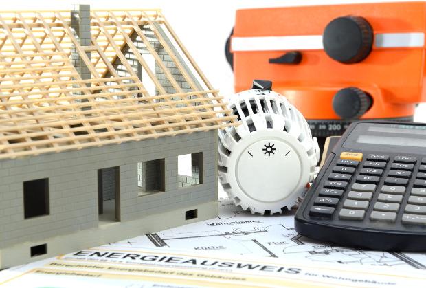 Im Bild zu sehen ist das Modell eines Neubaus, neben einem Thermostaten und einem Taschenrechner Heizung sanieren